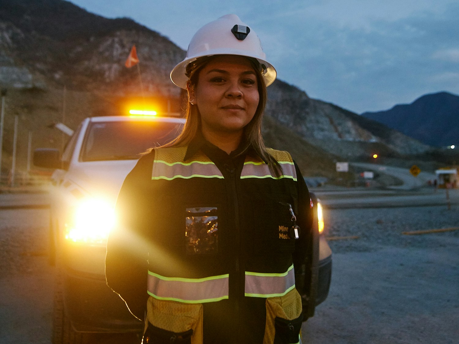 A woman wearing hazard clothing in front of truck with its lights on.